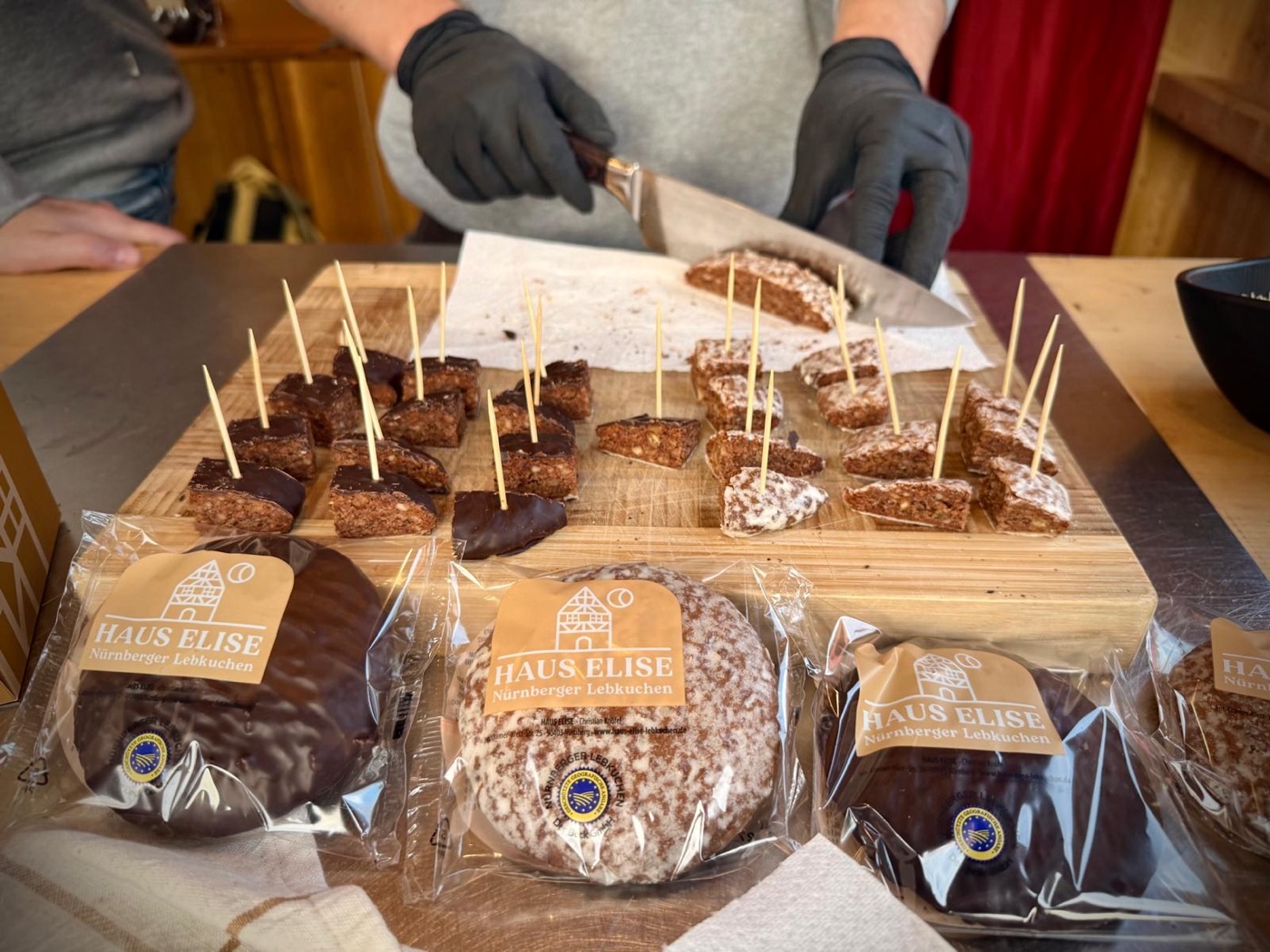Aufgeschnittene Elisen-Lebkuchen als Probierst&uuml;cke mit Holzspie&szlig;en auf einem Brett, davor einzeln verpackte Lebkuchen mit g.g.A.-Siegel